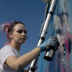 Woman creating vibrant graffiti art on a wall using spray paint while standing on a ladder.